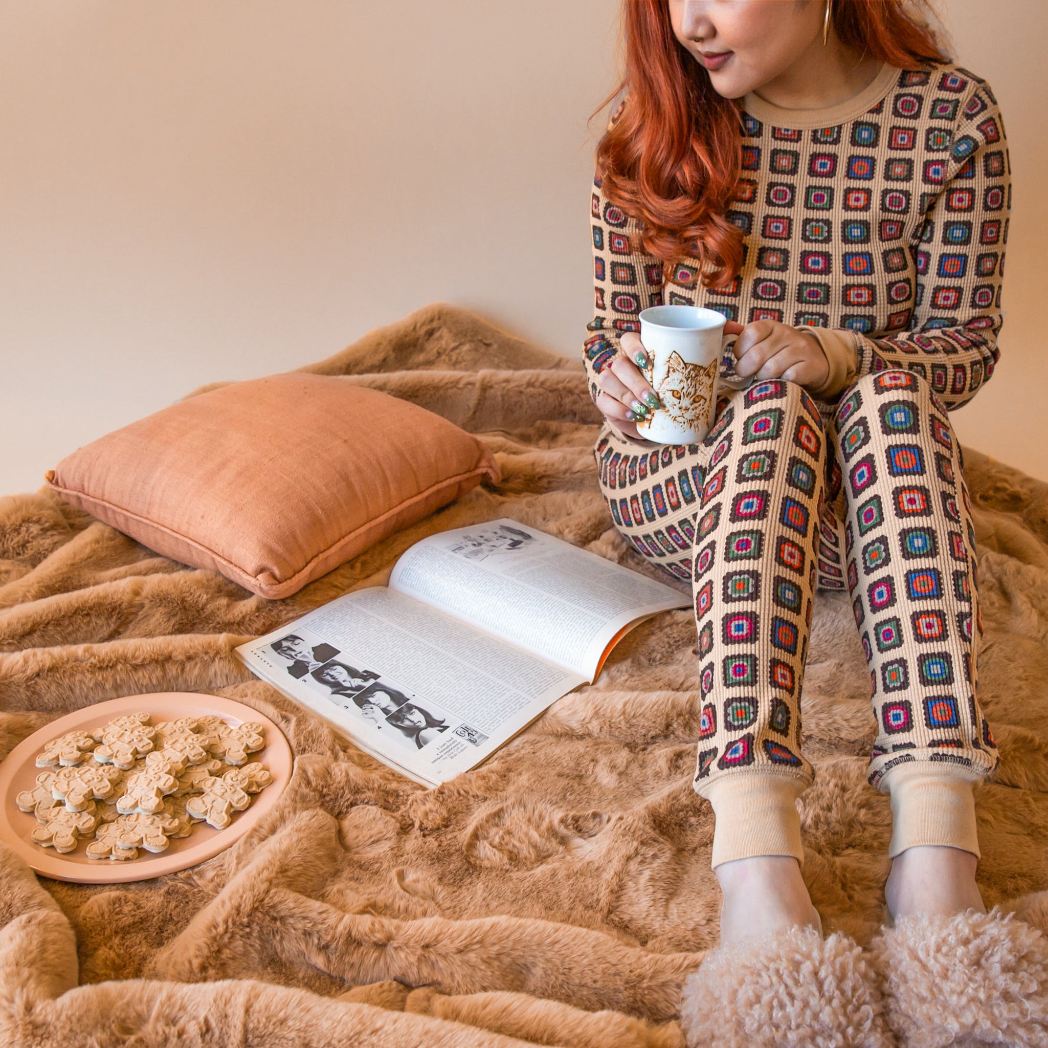 A woman wearing a matching thermal pj set sits on a brown fur blanket looking at a magazine holding a mug with a cat on it.