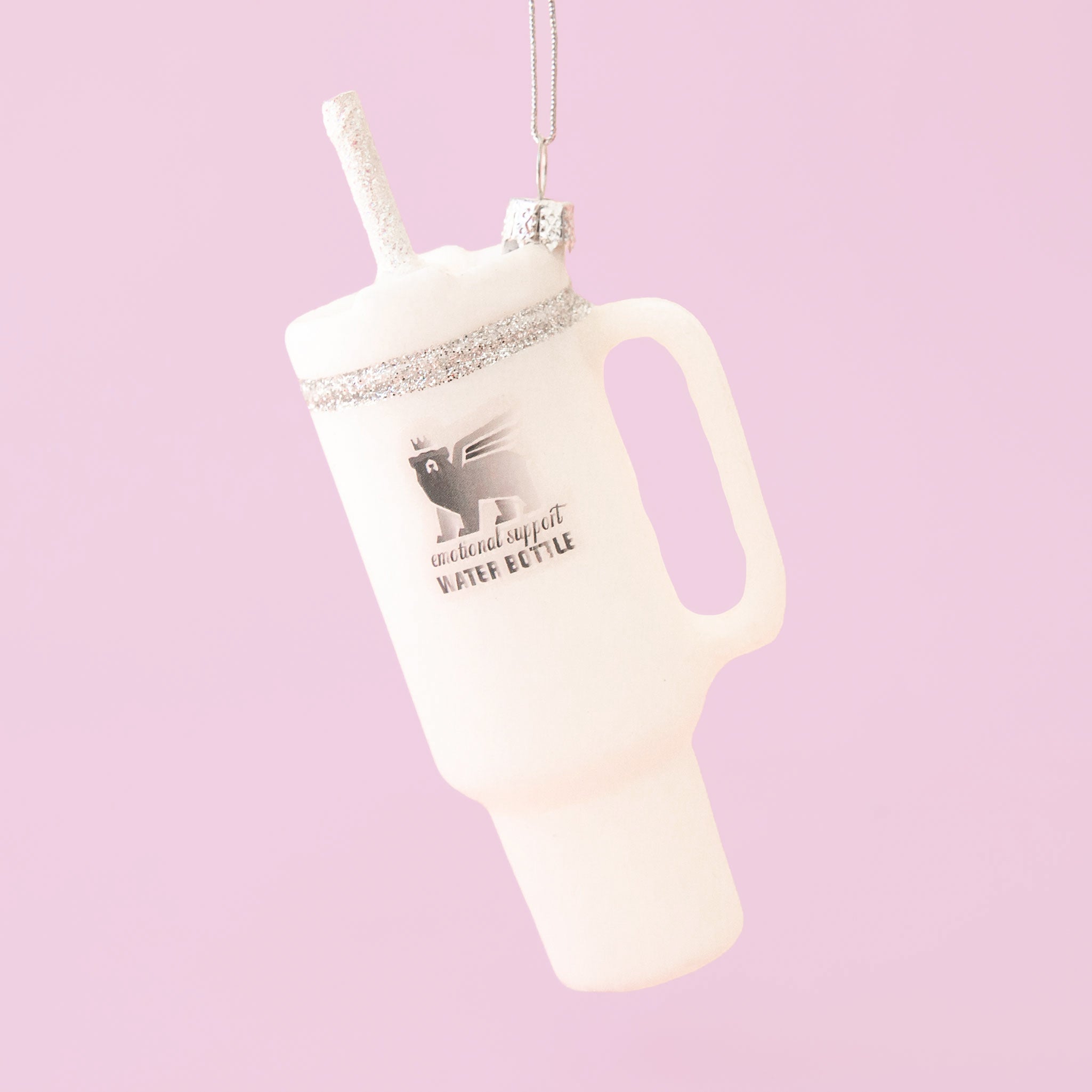 A white drinking tumbler ornament shown against a pink background. The lid and straw have silver sparkle details. 
