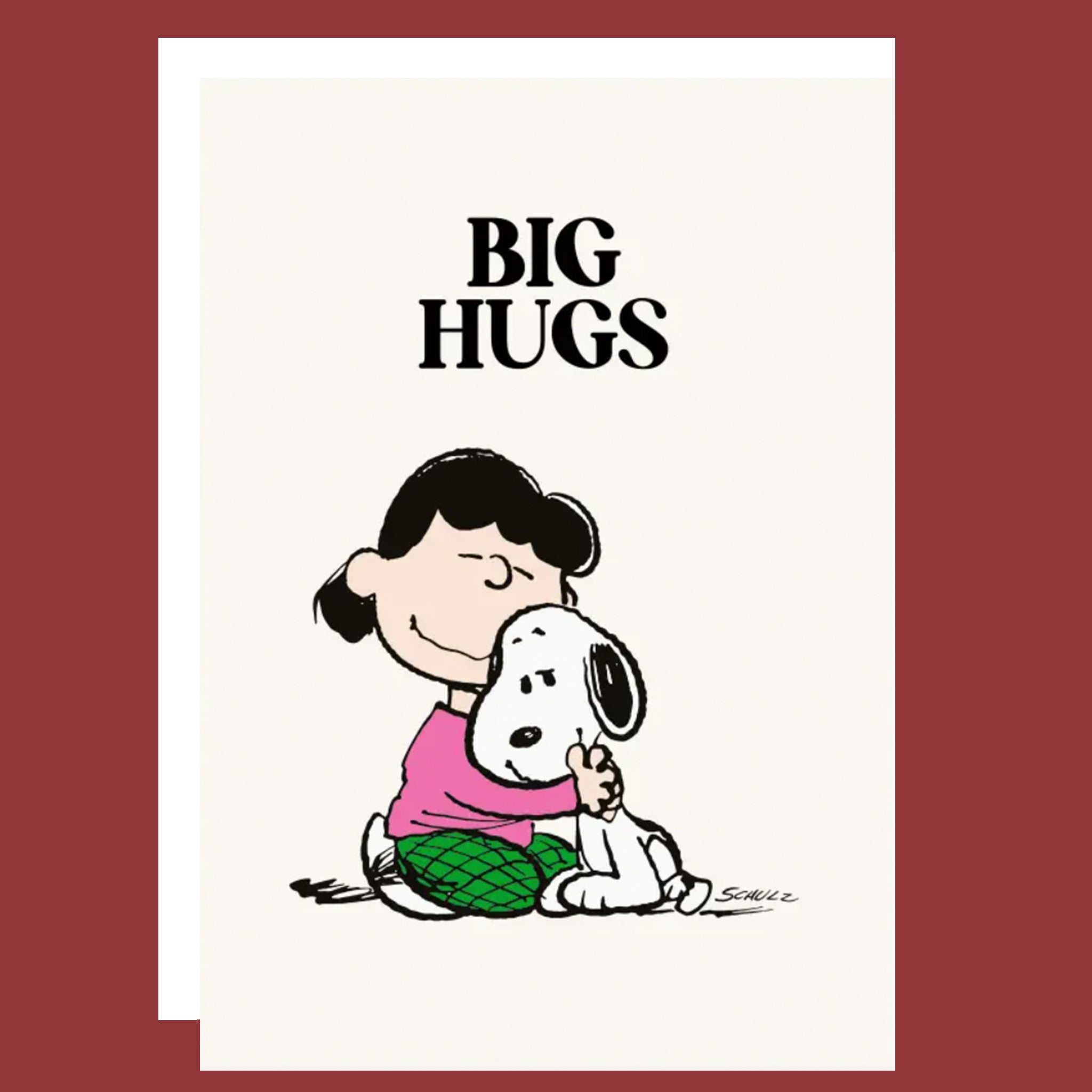 A greeting card featuring the classic Peanuts illustration of Lucy hugging Snoopy, with the text 'BIG HUGS' above them. The card has an off-white background, shown layered over another card against a solid dark red background.
