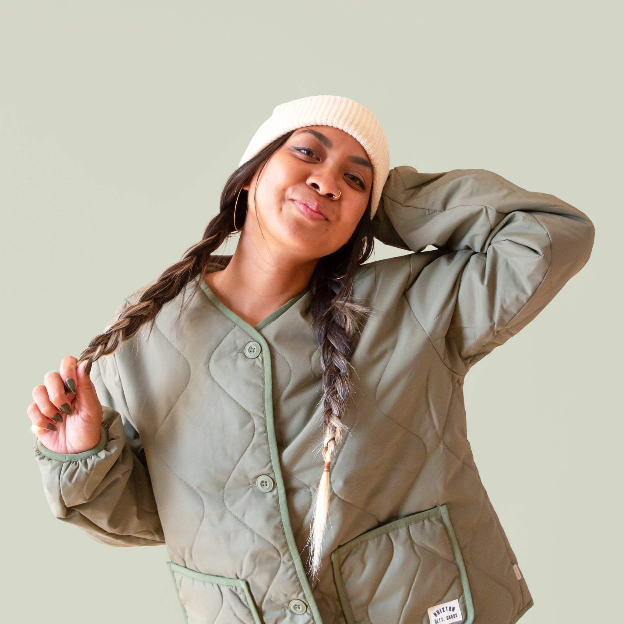 A girl wears an army green colored jacket, braids and a cream colored knit beanie.