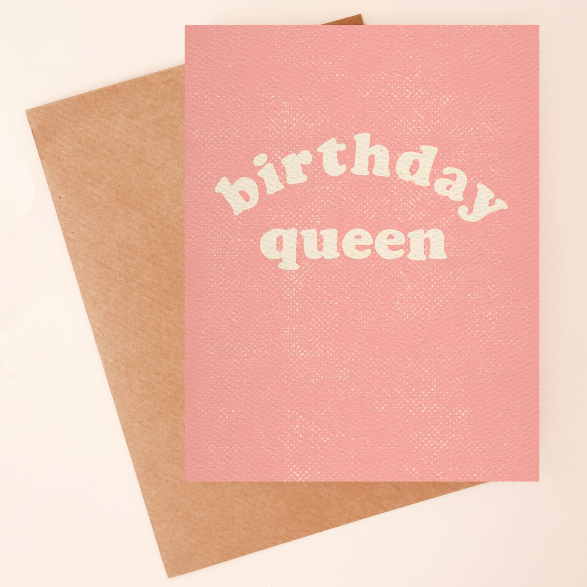 On a neutral background is a pink birthday card with a Kraft brown envelope that says, "birthday queen" in white text.
