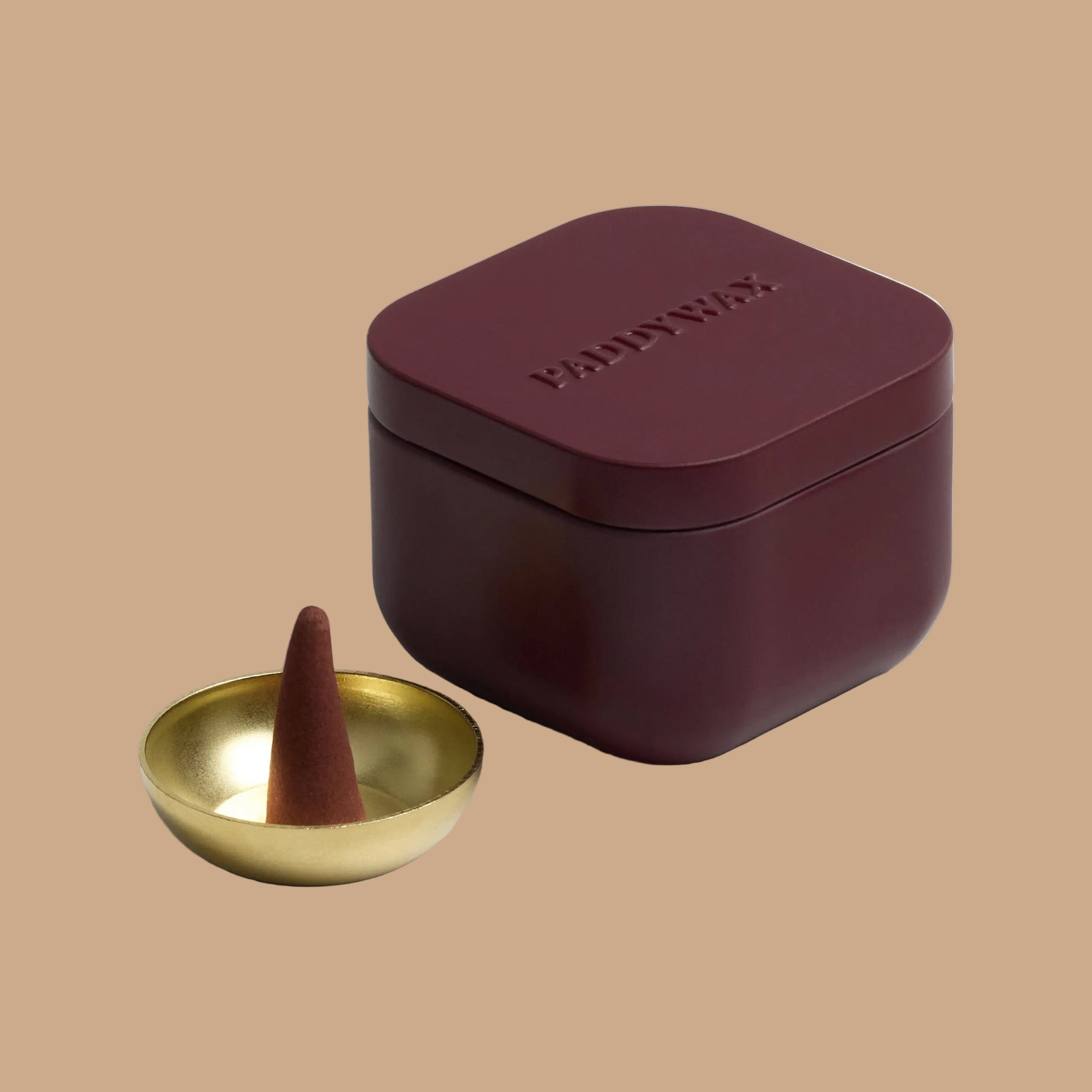 A burgundy tin filled with burgundy cone shaped incense and a gold tiny bowl for burning. 