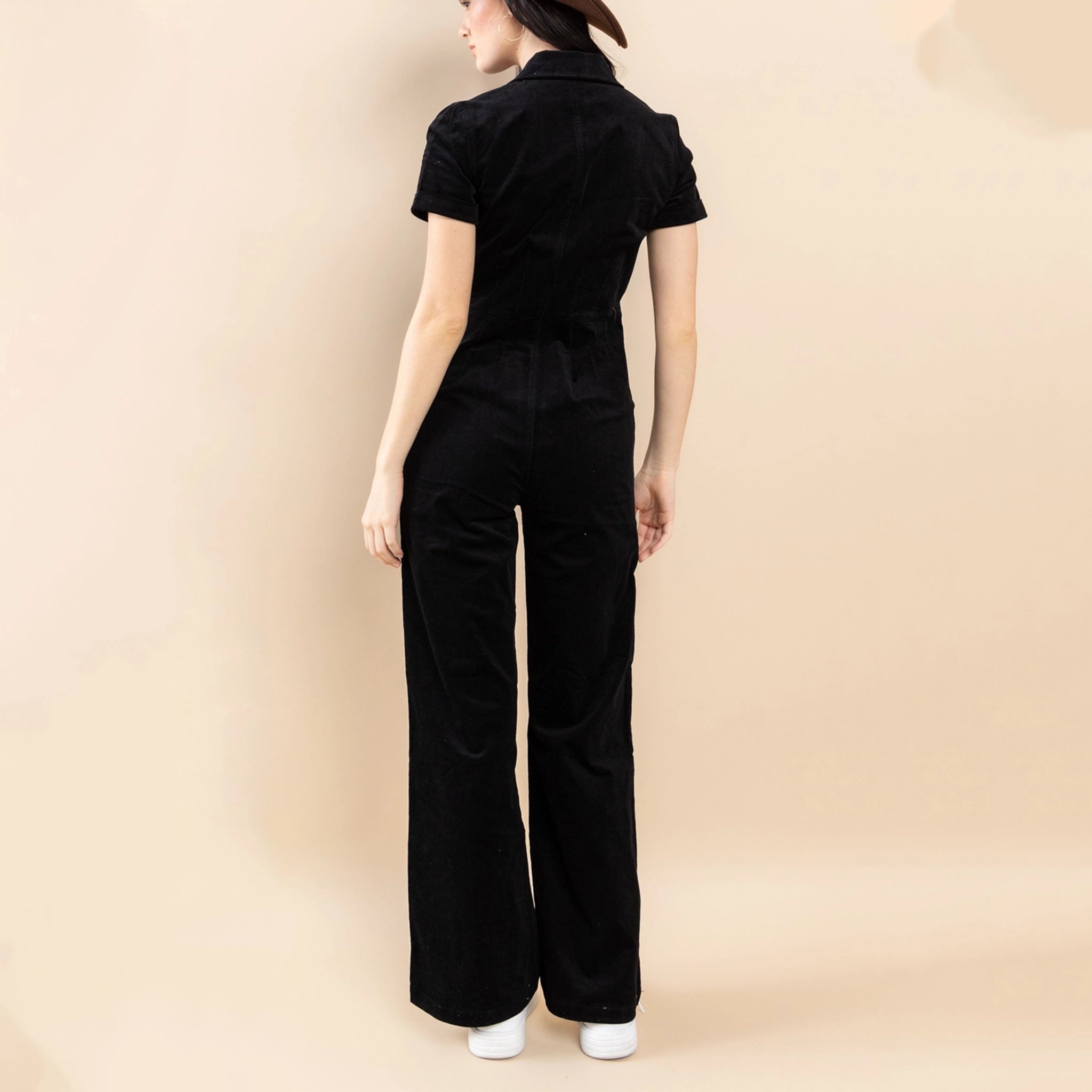 On a neutral background is a model wearing a black short sleeve jumpsuit with a front zipper.