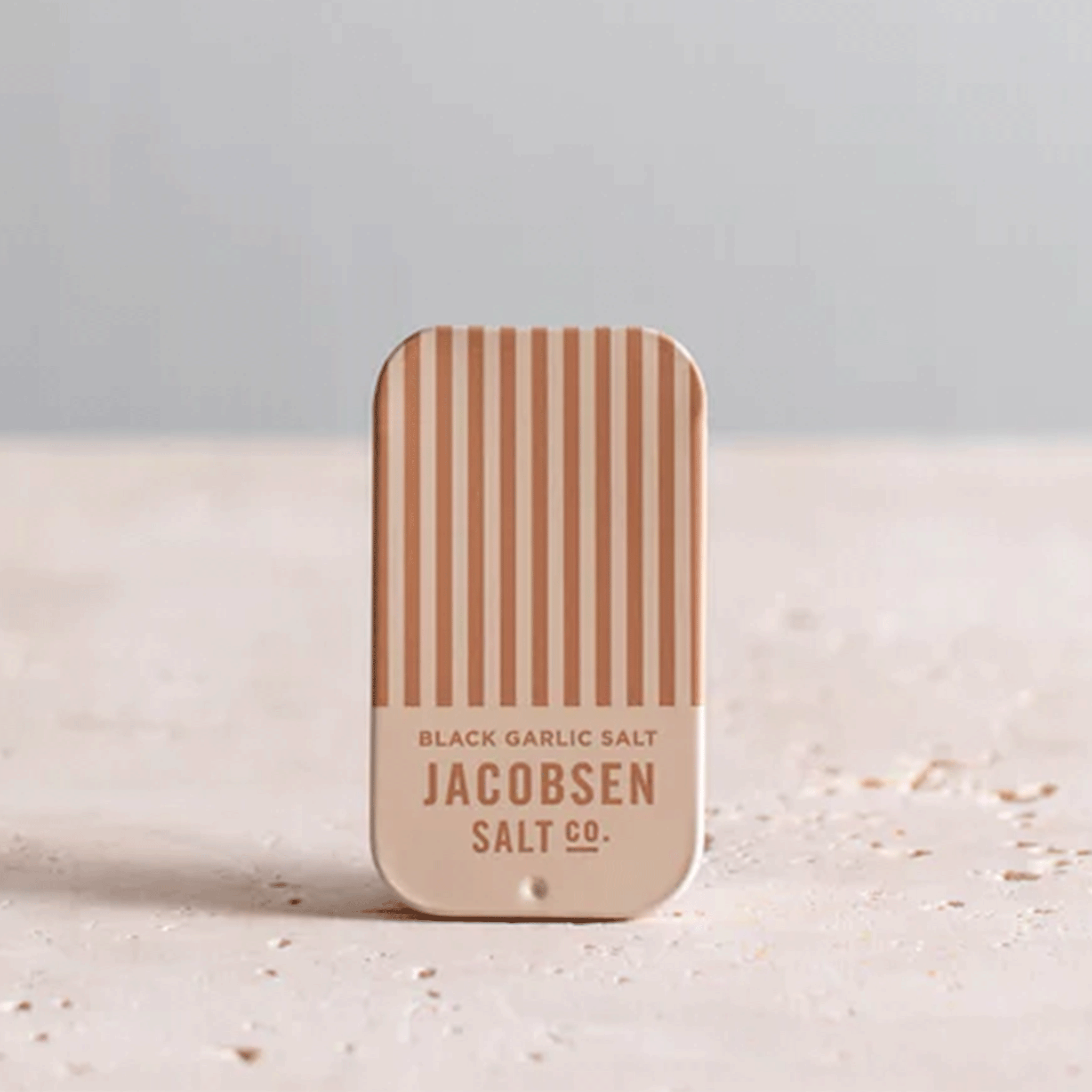A small tan and brown striped tin with text that reads, &quot;Black Garlic Salt Jacobsen Salt Co.&quot;.