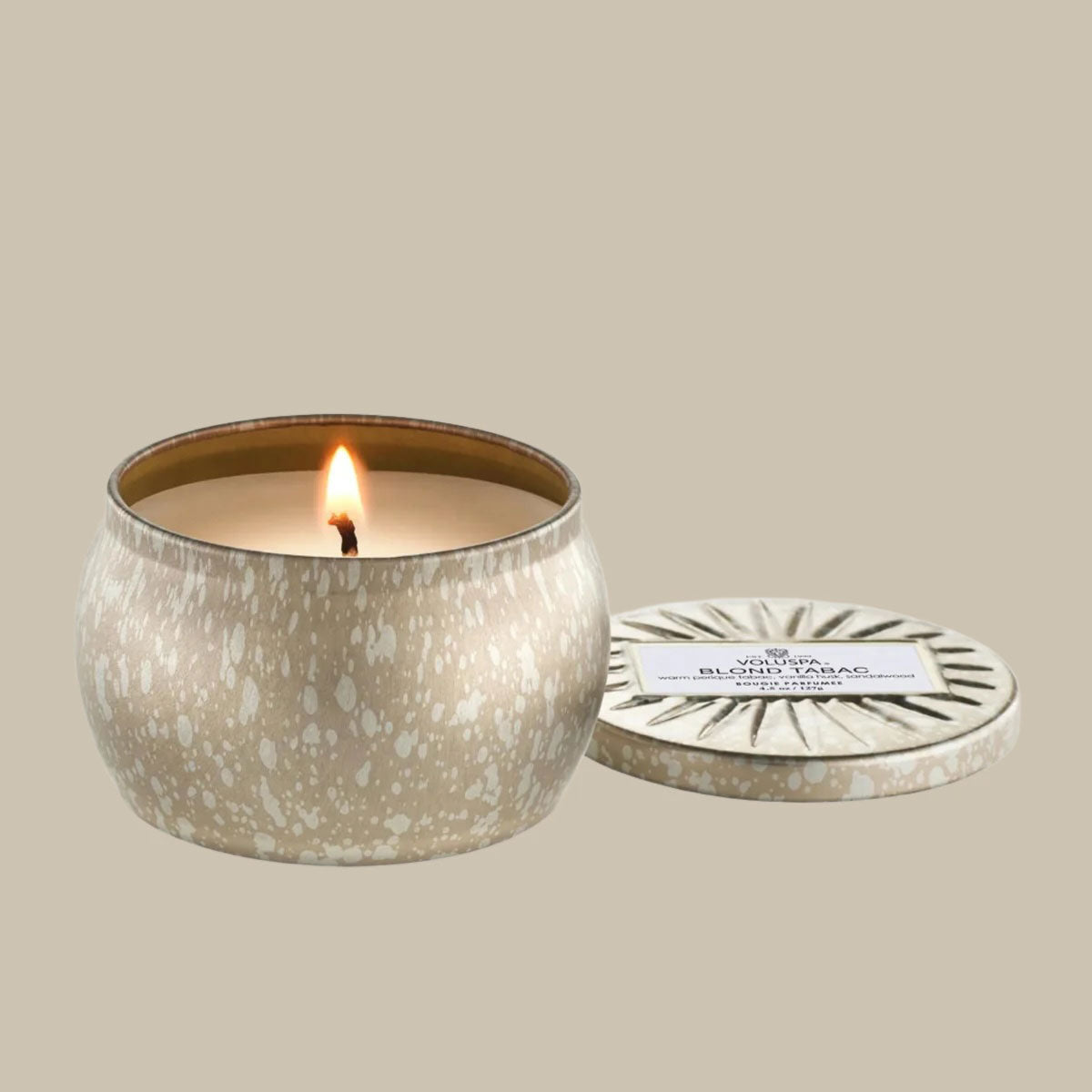 On a cream background is a light silver and gold mini tin candle with white wax and a single wick. Also included is the matching lid.