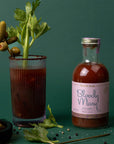 A glass bottle of bloody mary mix with a pink label and a gold lid. 