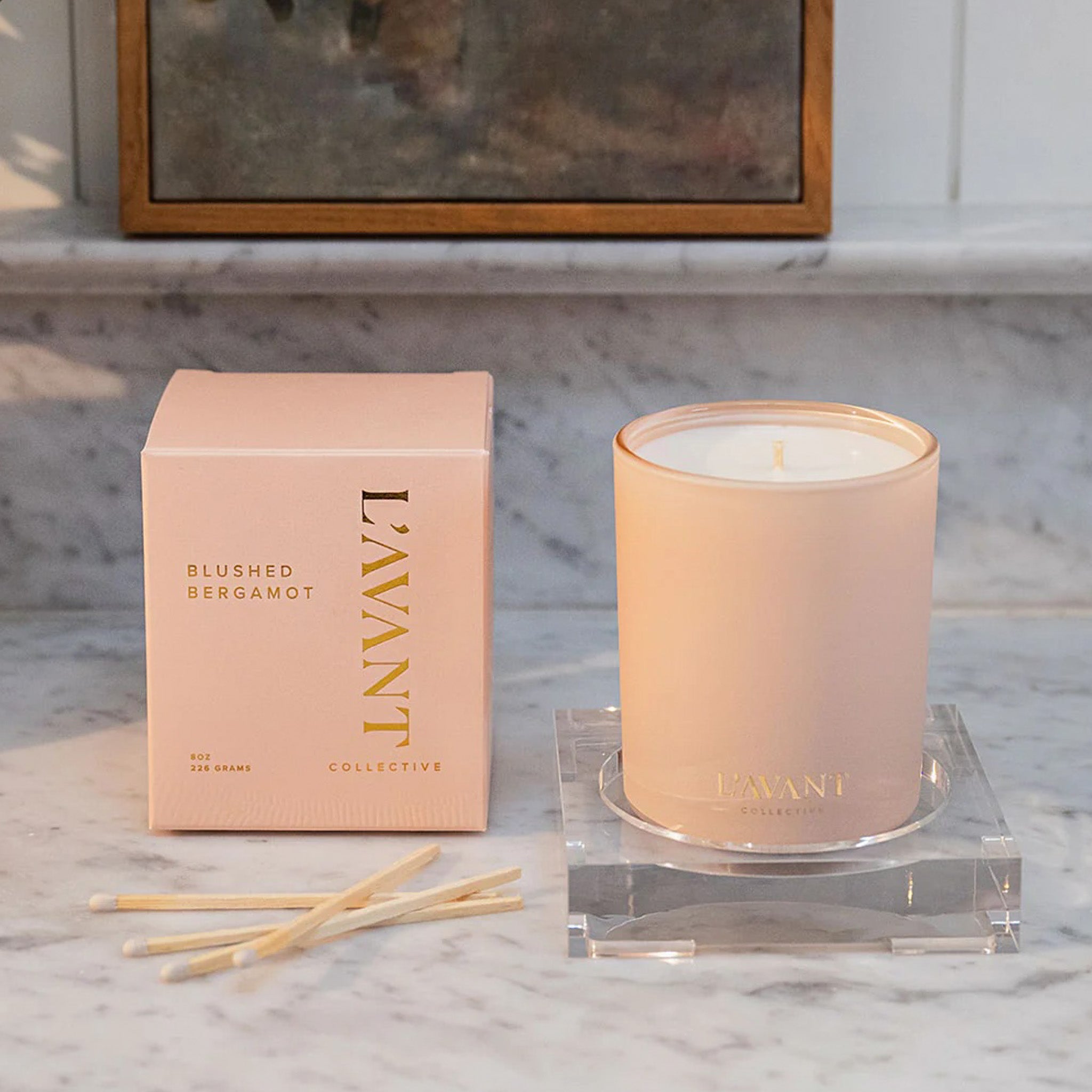 A pink candle on a marble counter seated next to the pink box it comes in.  The packaging reads "L'avant Blushed Bergamot"