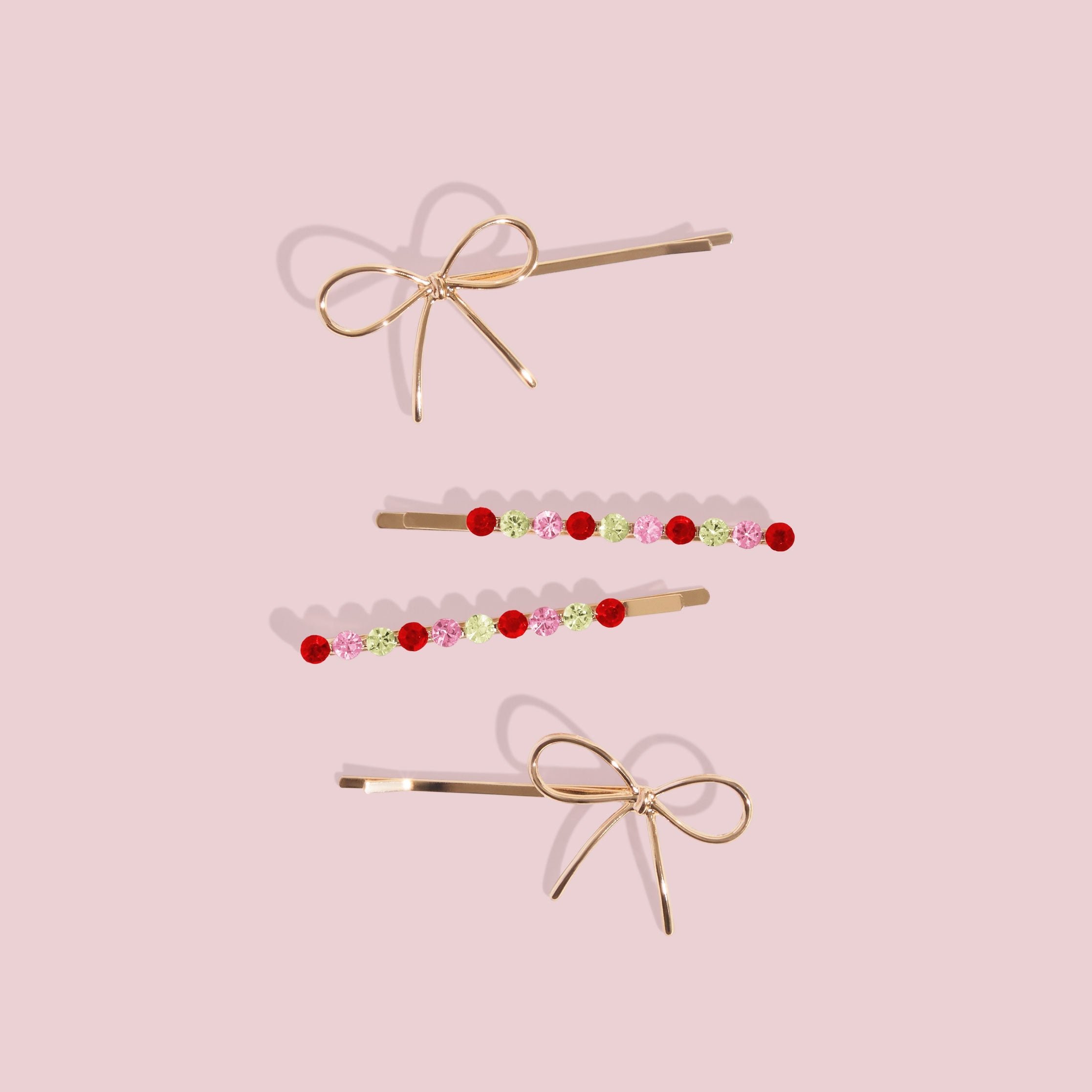 Two gold bow-shaped hair pins and two jeweled bobby pins with red, pink, and green stones arranged on a pale pink background.