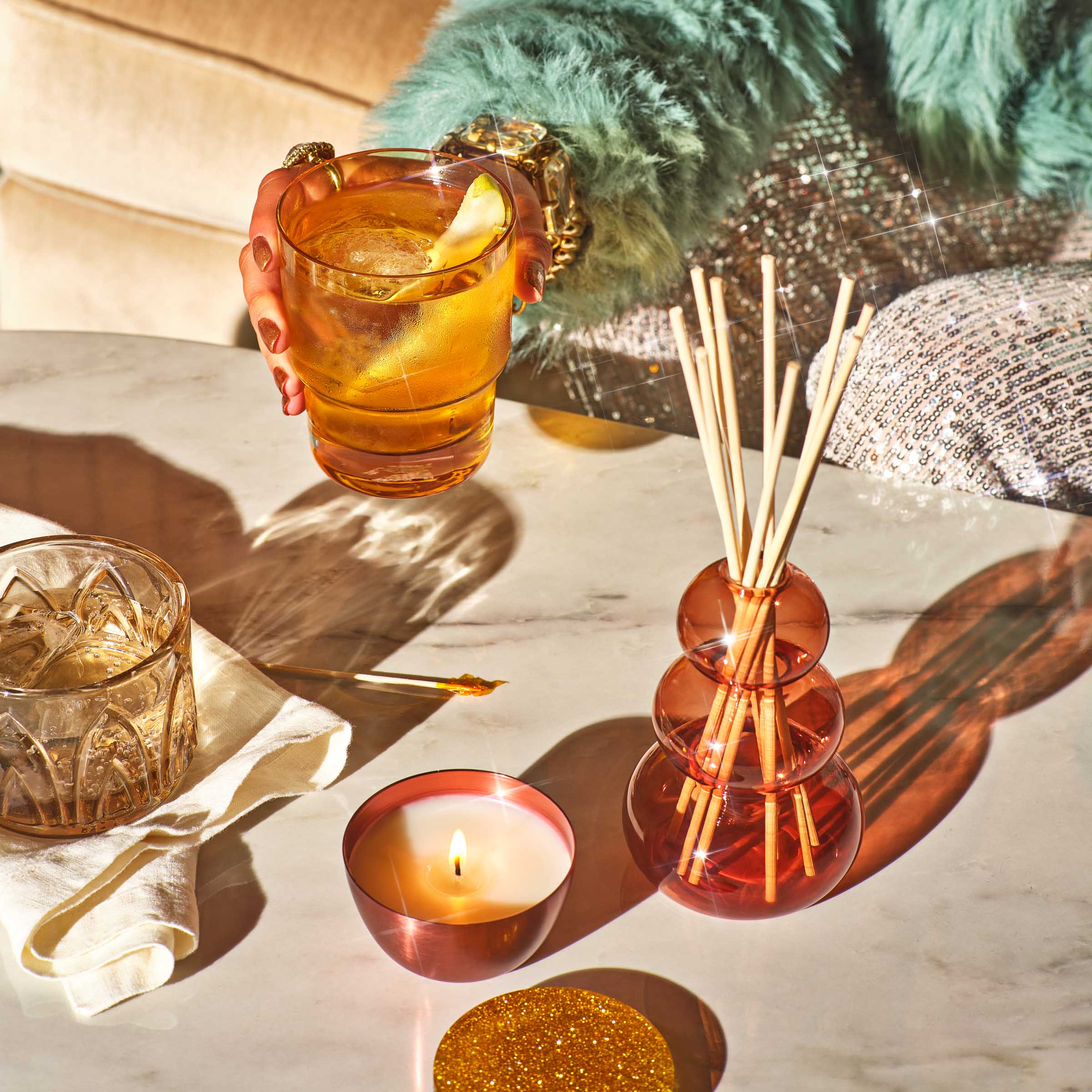 A hand holding a cocktail with a pear slice in it and amber glass vessels on the table in front.  A candle is lit in a jar with the lid covered in glitter.
