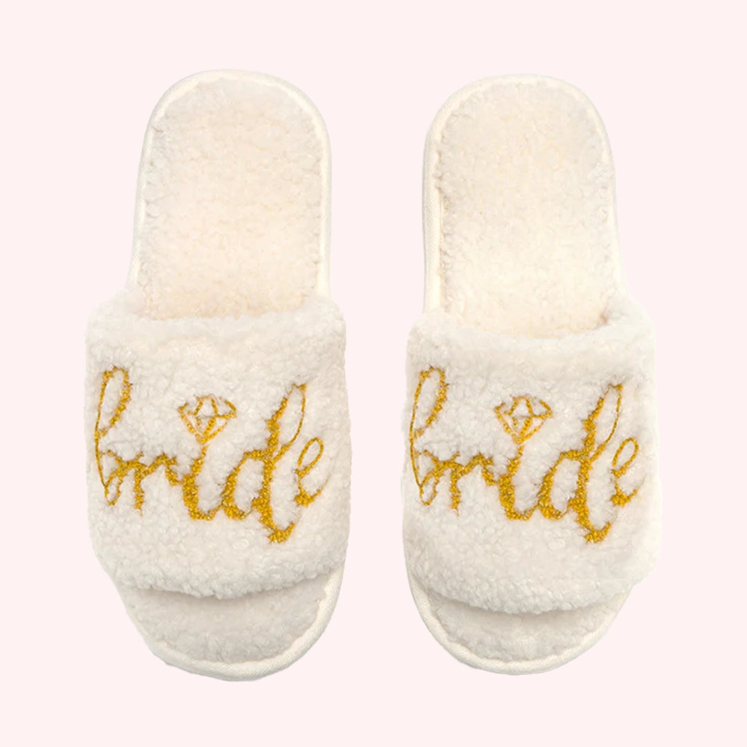 On a light pink background is a pair of fuzzy white bride slippers with gold writing on the front that reads, &quot;Bride&quot; with a diamond shape used above the &#39;i&#39;.