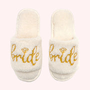 On a light pink background is a pair of fuzzy white bride slippers with gold writing on the front that reads, "Bride" with a diamond shape used above the 'i'.