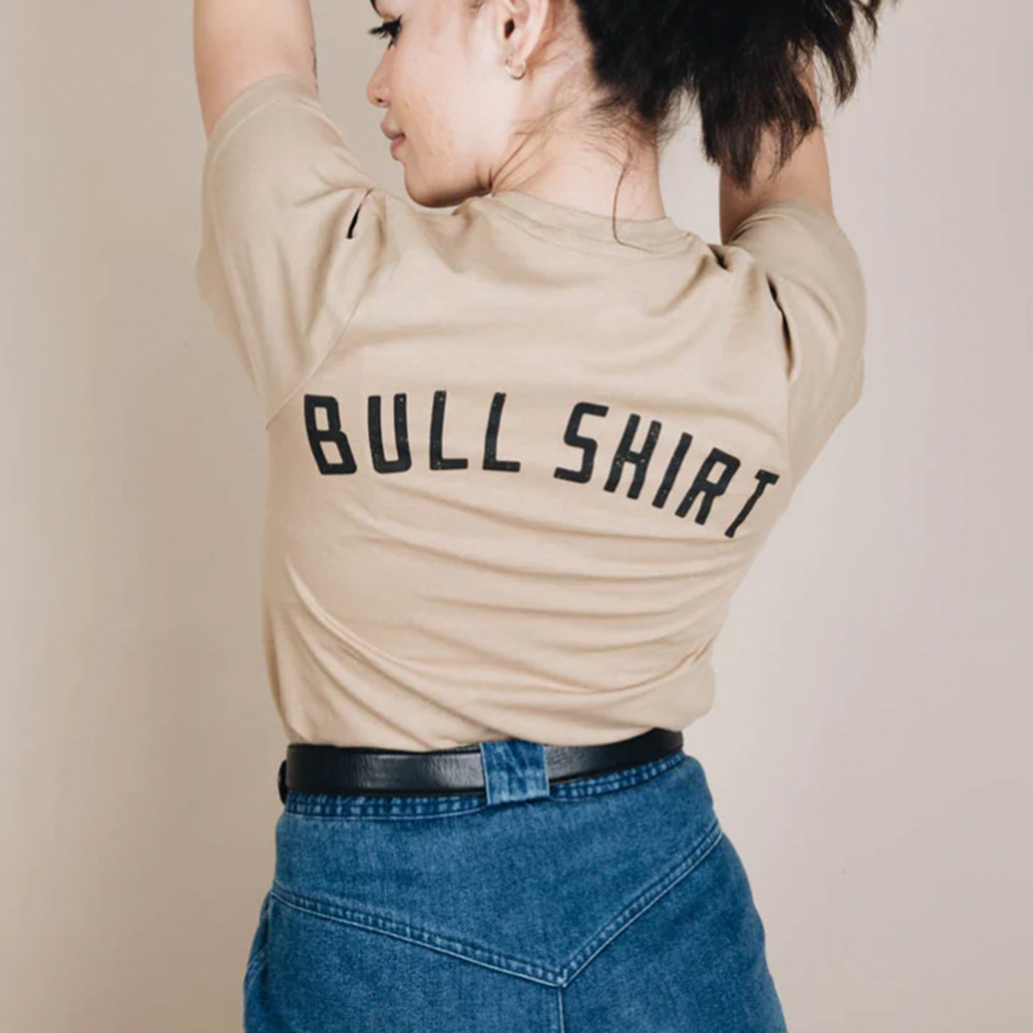 On a tan background is a tan t-shirt with a black bull screen printed on the front and text on the back that reads, "Bull Shirt".
