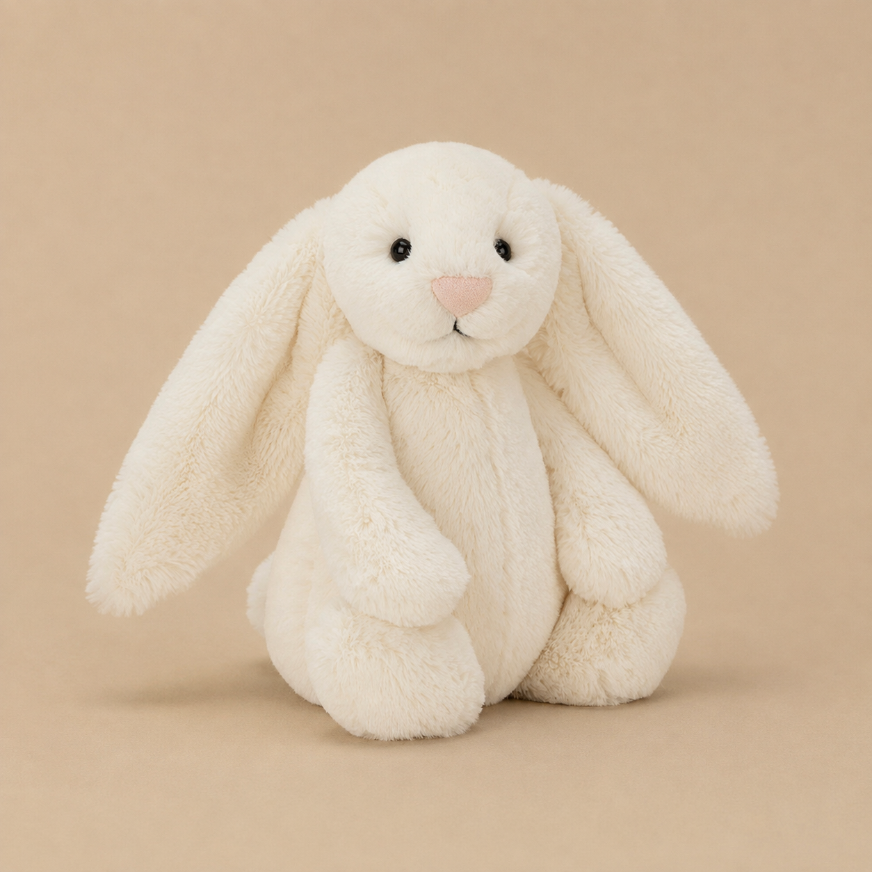 A plush cream-colored bunny toy with long floppy ears, a pink stitched nose, and small black eyes, photographed on a clean white background.