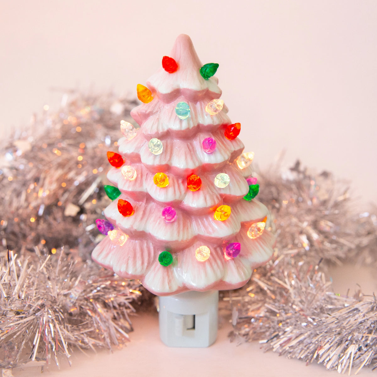 A retro-style pink ceramic Christmas tree light with multicolored bulb accents, set against a soft pink background with silver tinsel garland.