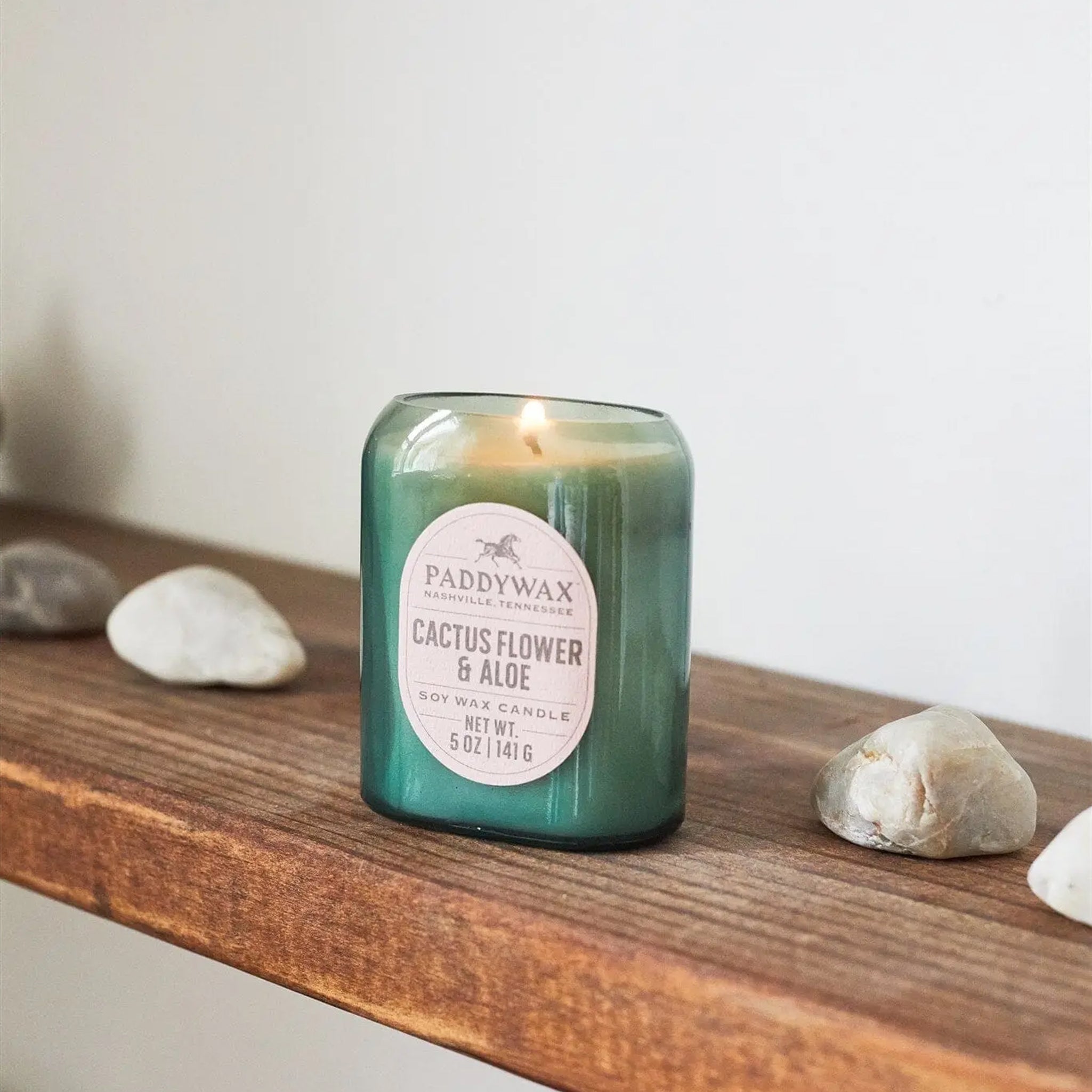 Green glass candle labeled ‘Cactus Flower & Aloe’ burning on a wooden shelf, with smooth decorative stones placed around it.