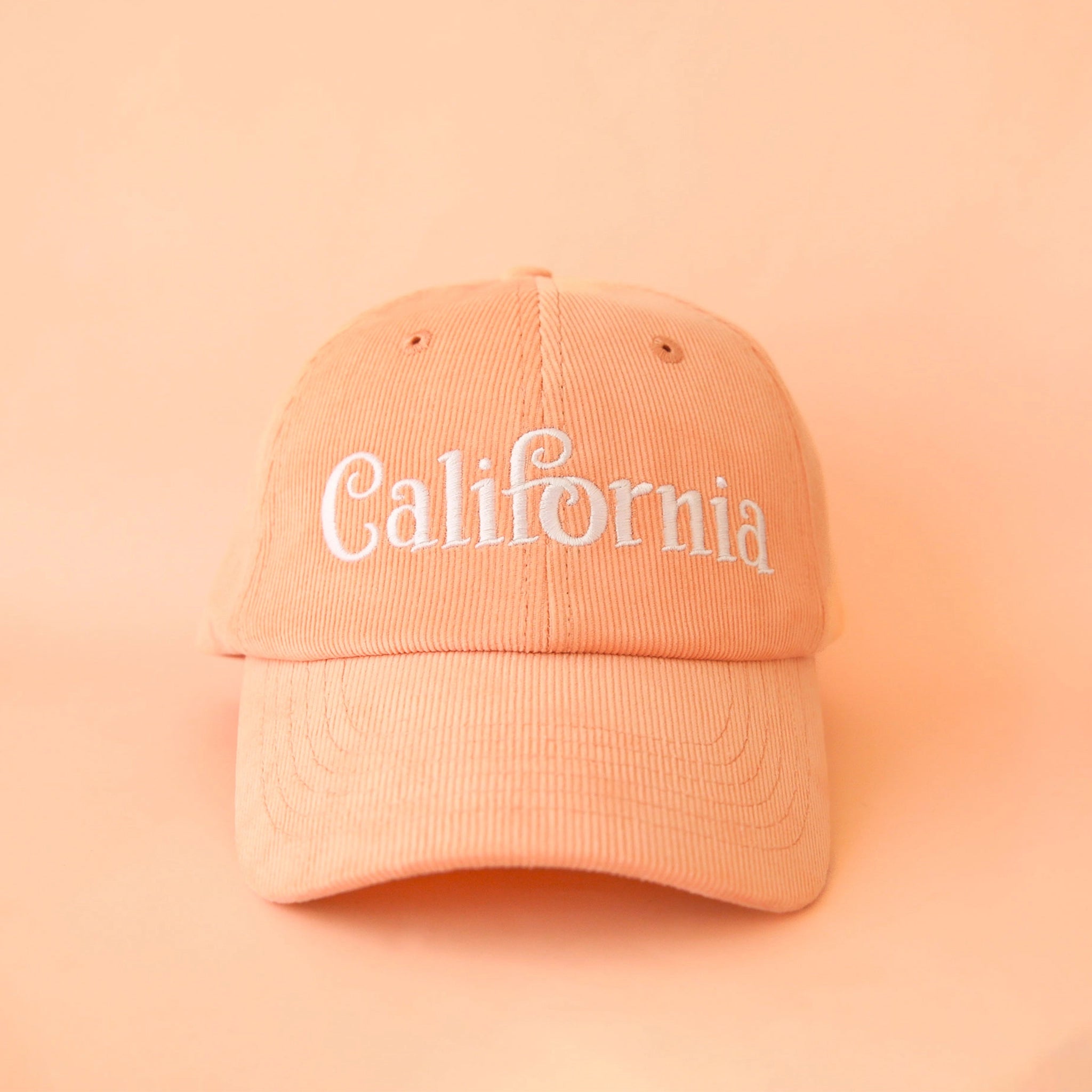On a peach background is a peach corduroy baseball hat with white embroidery that reads, "California".