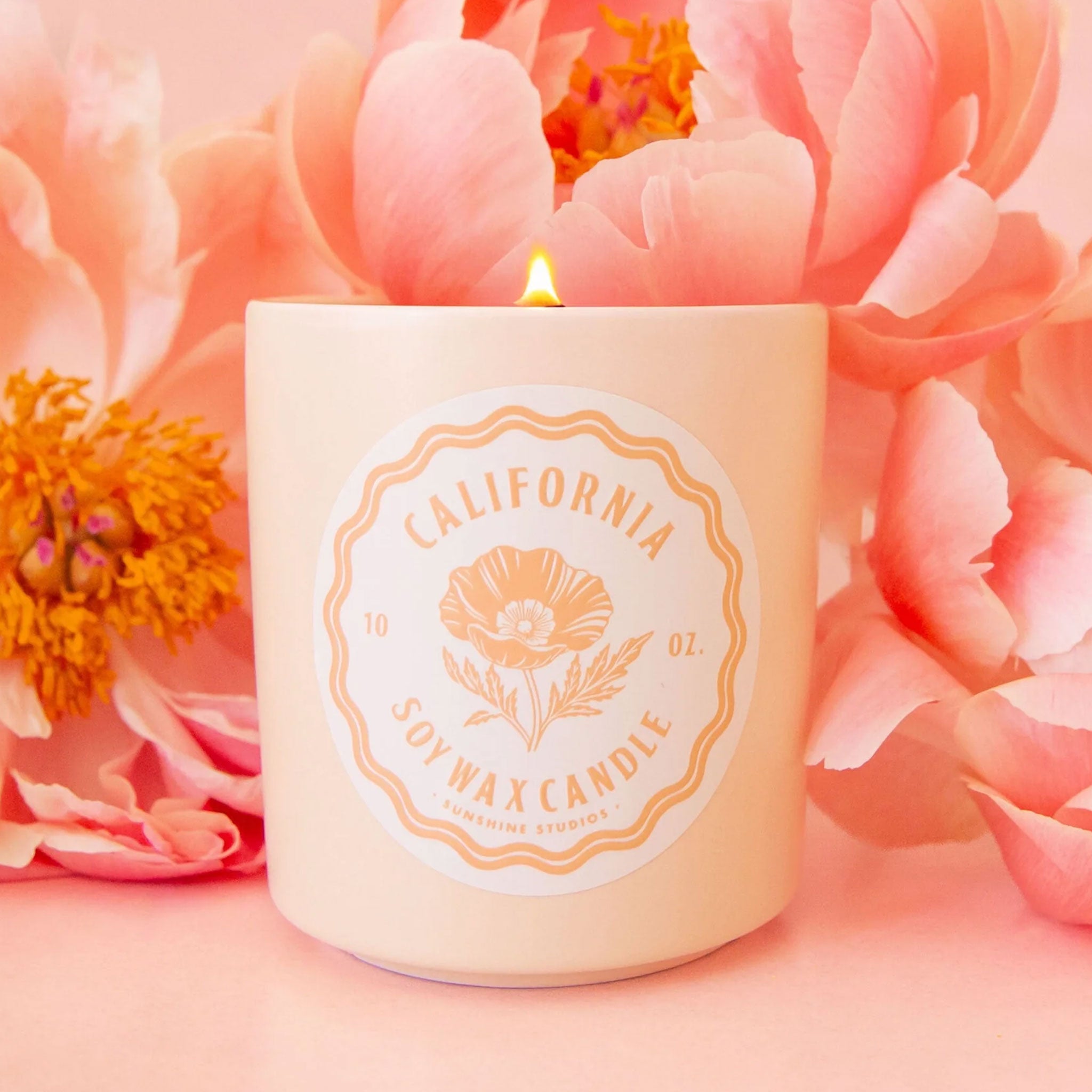A ceramic candle sits in the middle of image.  The candle has a round white label that reads, "California Soy Wax Candle", in hte background are pink peony flowers.