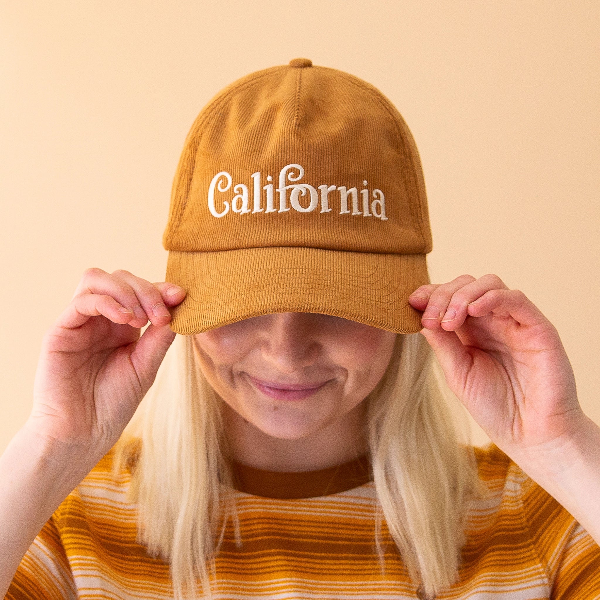 On a peachy background is a model wearing a toffee colored corduroy snapback hat that has white text embroidered across the front reading, &quot;California&quot;. 