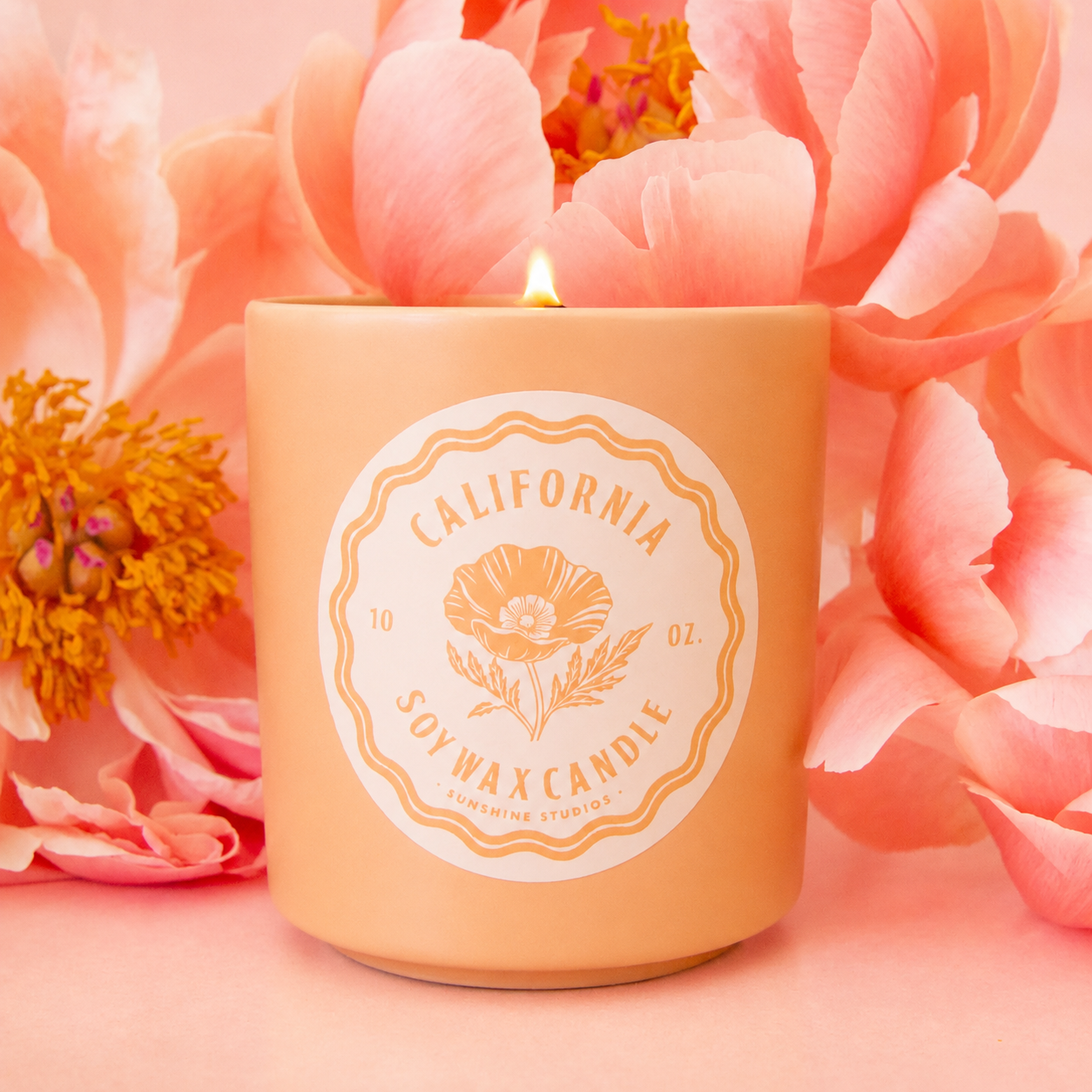 A ceramic candle sits in the middle of image.  The candle has a round white label that reads, "California Soy Wax Candle", in hte background are pink peony flowers.