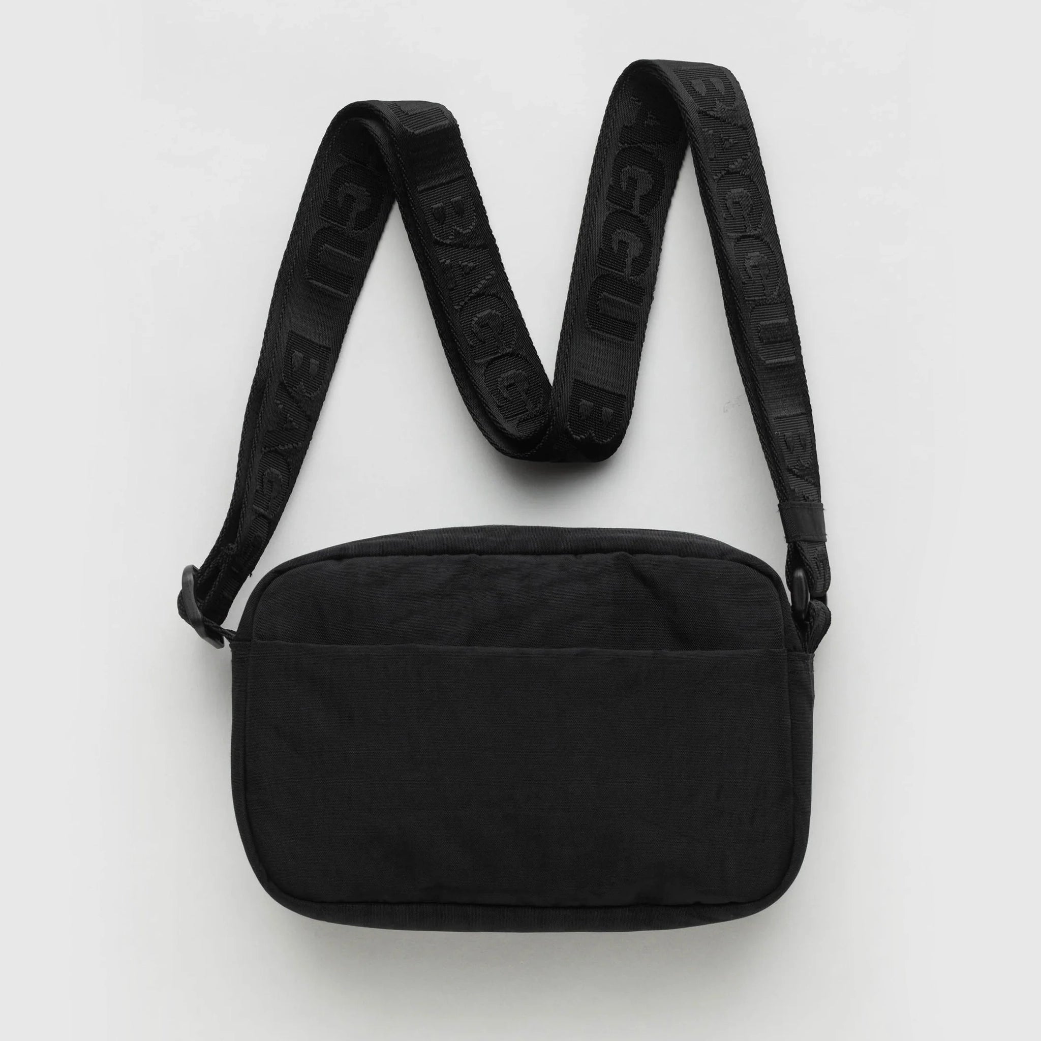On a light grey background is a black crossbody made of sturdy nylon material with a black adjustable strap that has &quot;BAGGU&quot; stitched into it in a shinier black material.