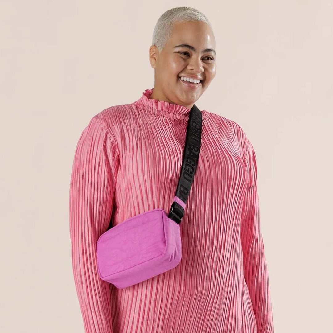 On an ivory background is a bright pink crossbody made of sturdy nylon material with a black adjustable strap that has &quot;BAGGU&quot; stitched into it in a shinier black material.