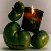 A green glass candle with small text in the front that reads, "Roma Heirloom Tomato Formulated for Daily Pleasure" surrounded by green tomatoes.