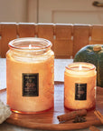 A large decorative jarred candle in an orange shade next to the smaller version with a matching lid and black label in the center that reads, "Voluspa Spiced Pumpkin Latte".