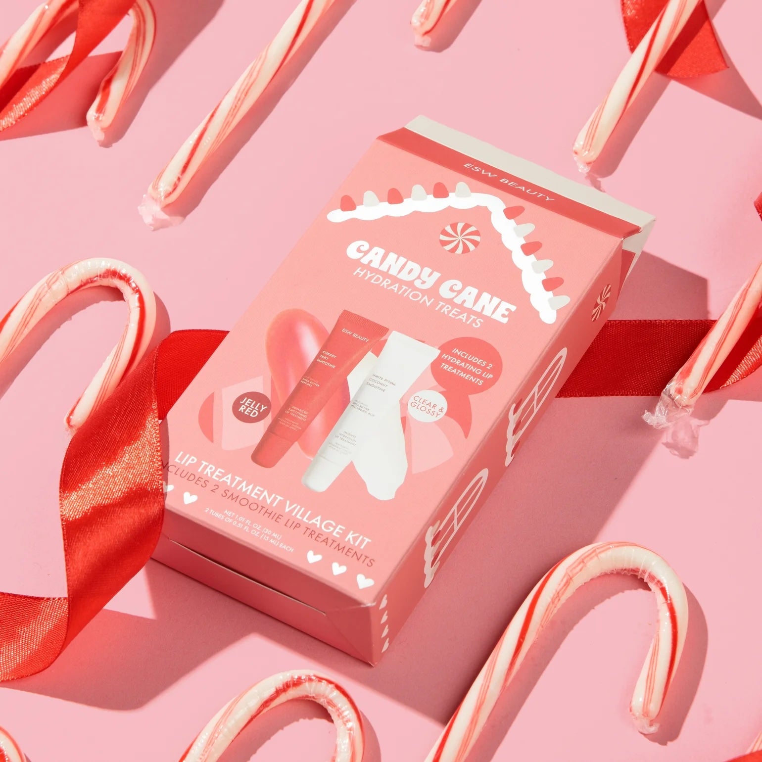 Pink ‘Candy Cane Hydration Treats’ lip treatment kit box on a pastel pink background, surrounded by candy canes and a red satin ribbon in a holiday flat-lay.