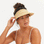 A hand woven what straw visor in a light blonde color. It features an elastic strap in the back that has slight give to fit most. This photograph shows a model wearing the visor.