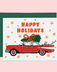 A white, red and green holiday card and envelope with a graphic of a vintage red car with a tree on top and text that reads, 'Happy Holidays'. 