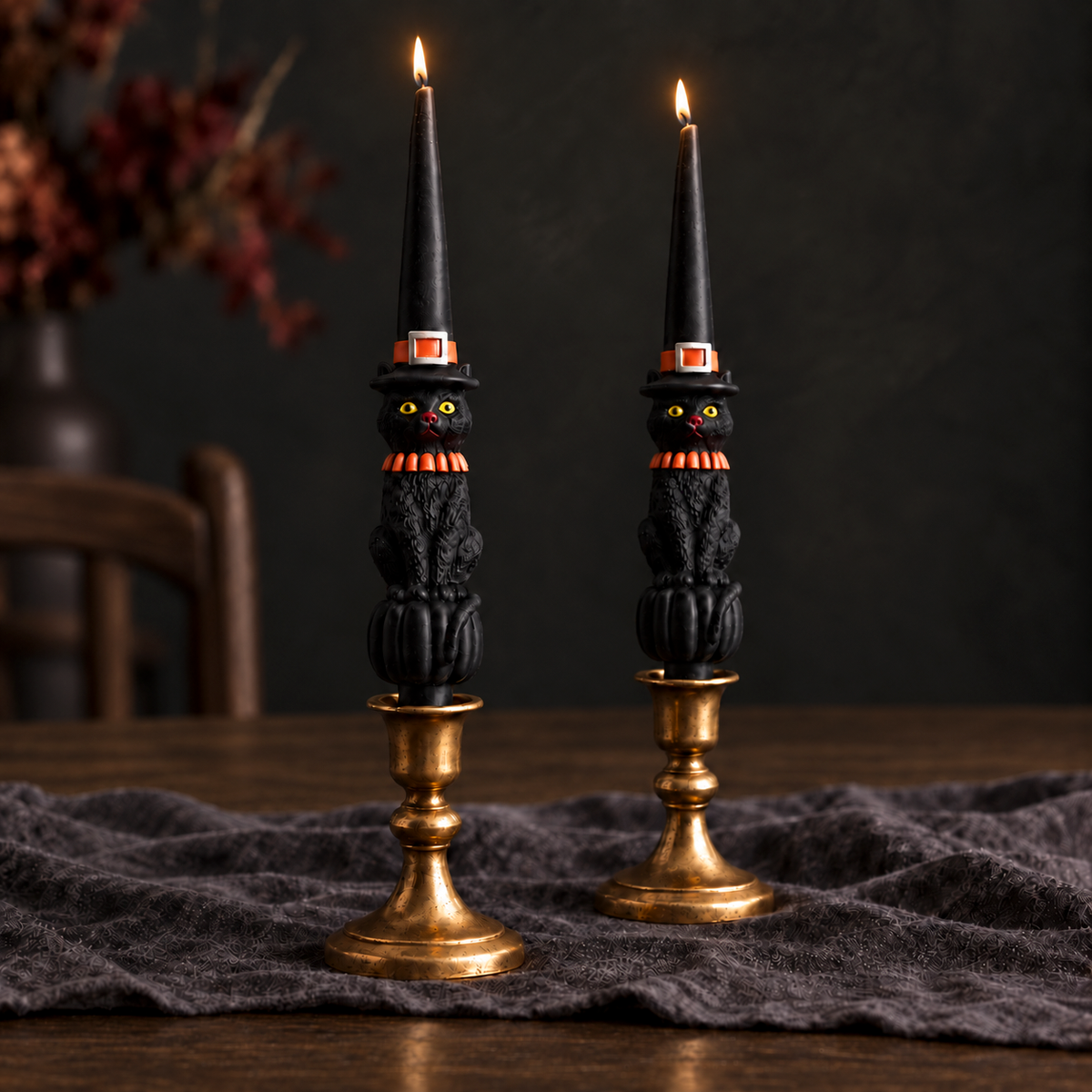 Black cat shaped taper candles in gold taper holders on a wooden table with a black cloth and black walls in background.