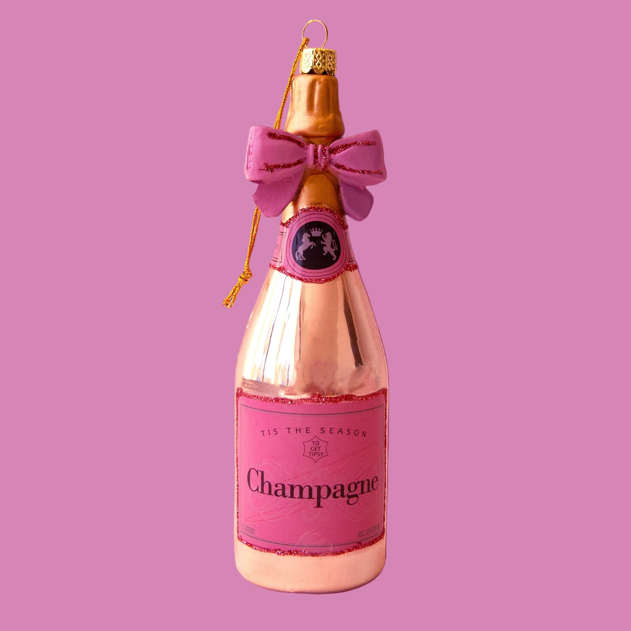 A rose gold champagne bottle shaped ornament with a pink label that reads, 'Tis The Season Champagne'. 
