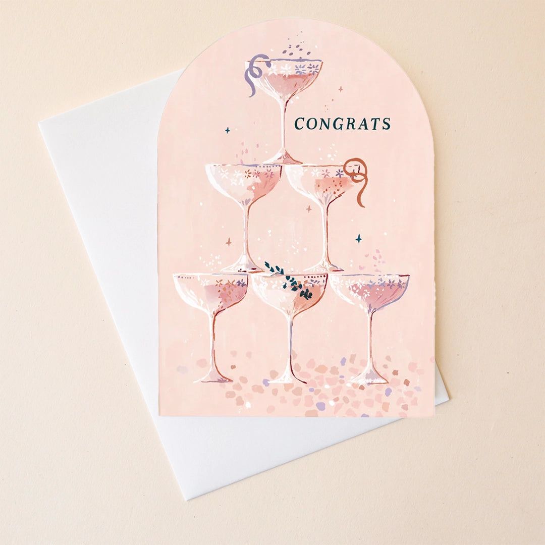 On a neutral background is a pink arch shape card that has illustrations of champagne coop glasses and text at the top that reads, "Congrats". 