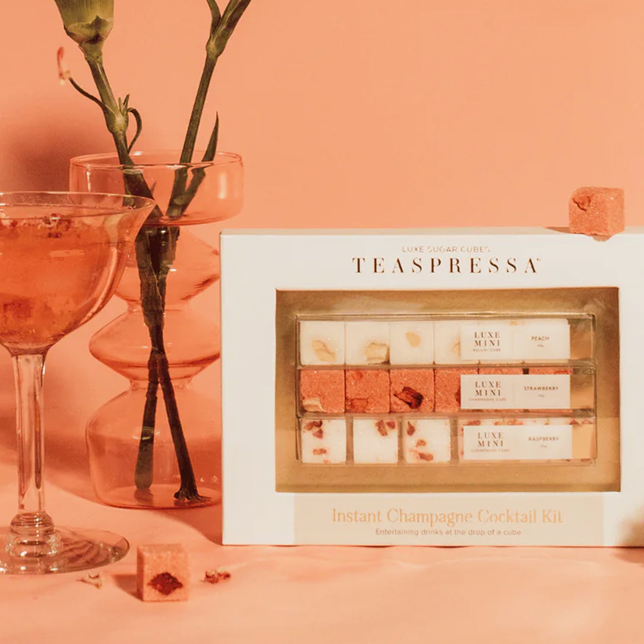 On a pink background is a sugar cube cocktail kit that reads, "Teaspressa Instant Champagne Cocktail Kit" with a set of three sugar cube strips that come in three flavors.