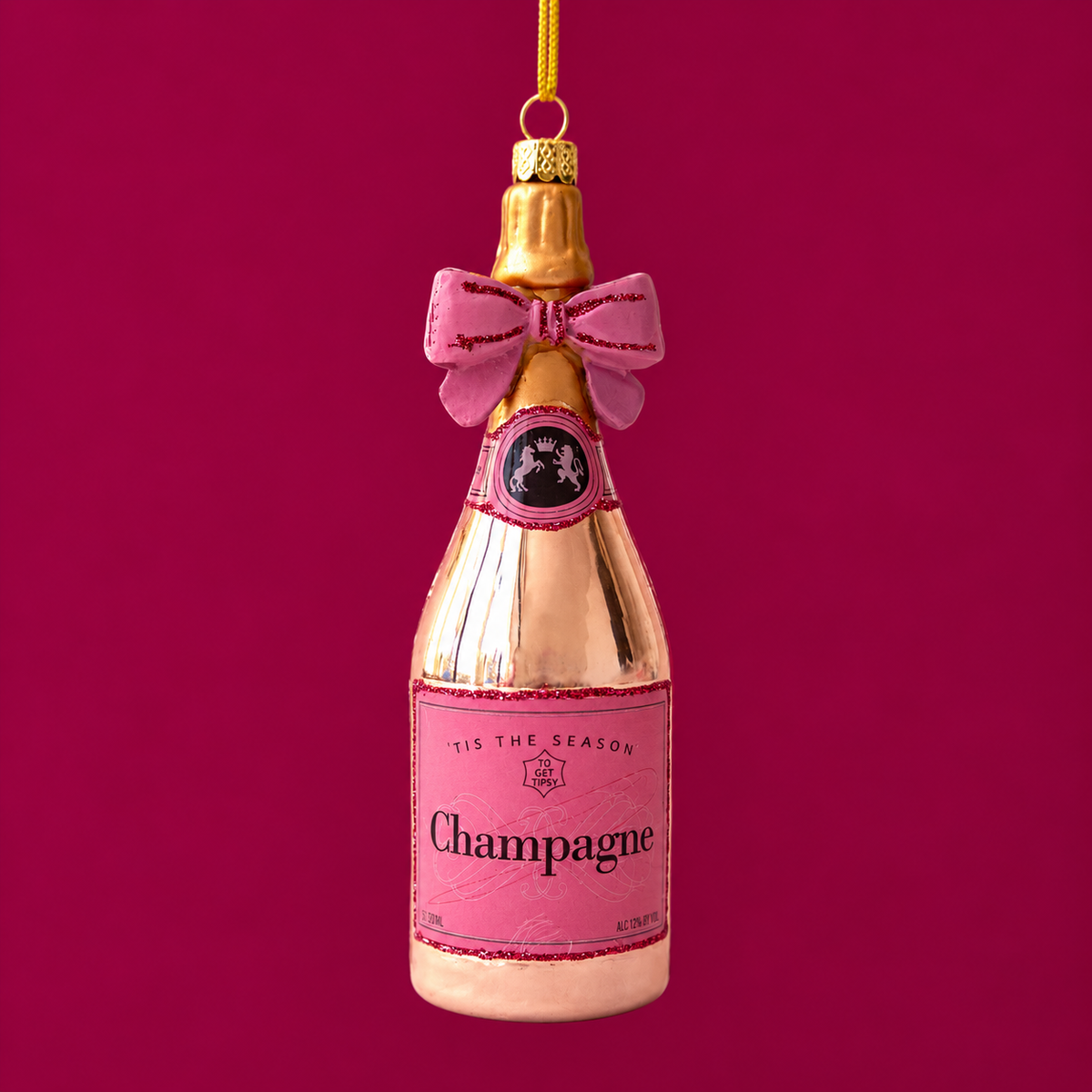 A rose gold champagne bottle shaped ornament with a pink label that reads, 'Tis The Season Champagne'. 