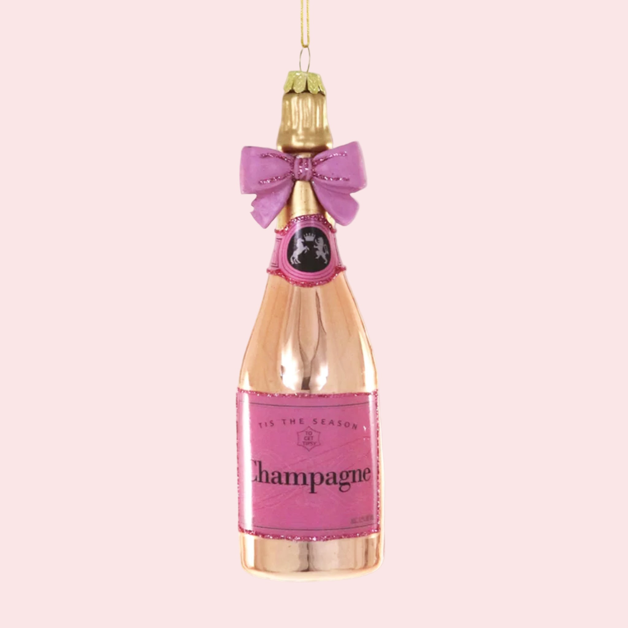 A rose gold champagne bottle shaped ornament with a pink label that reads, 'Tis The Season Champagne'. 