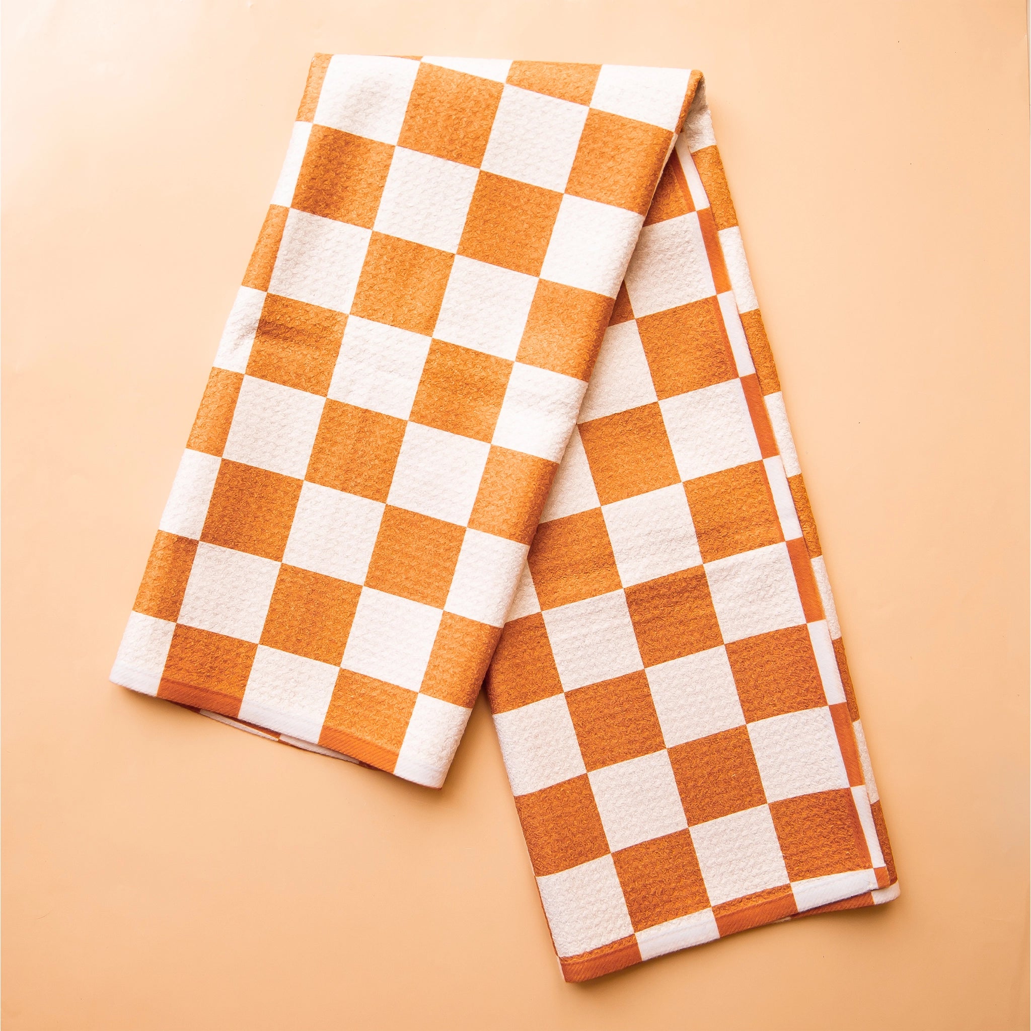 On an orange backdrop is an orange and ivory checkered kitchen towel.