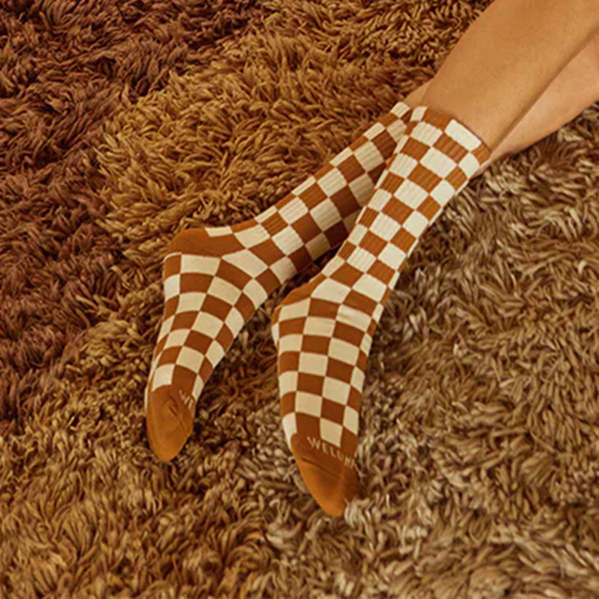 On a rust shag colored carpet is a model wearing a pair of rust and cream checker printed socks. 