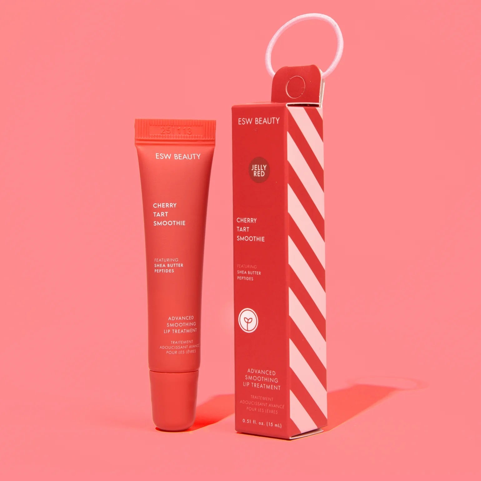 Red ESW Beauty ‘Cherry Tart Smoothie’ lip treatment tube standing next to its candy-cane striped red box on a bright pink background.