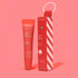 Red ESW Beauty ‘Cherry Tart Smoothie’ lip treatment tube standing next to its candy-cane striped red box on a bright pink background.