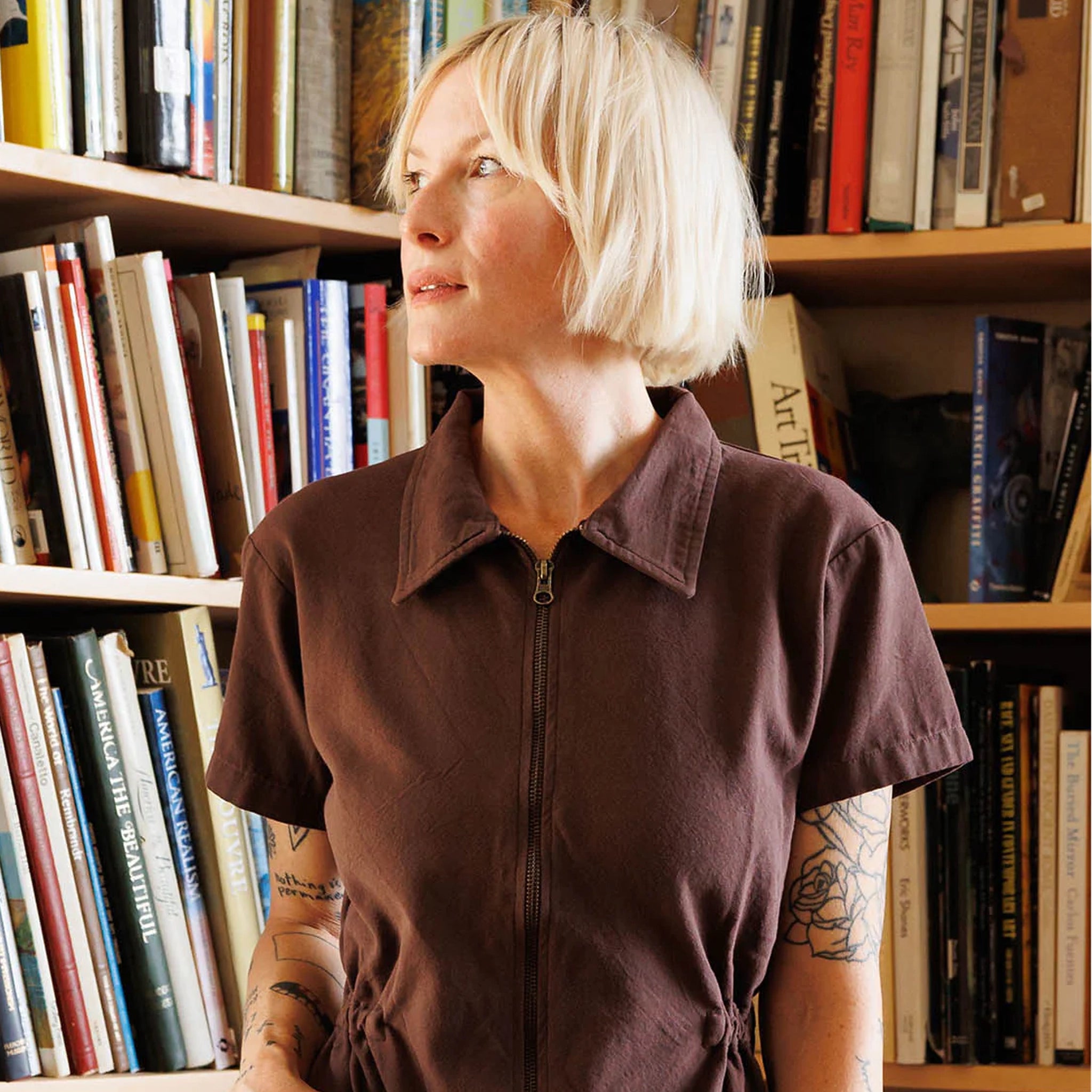A model standing in front of a bookcase with a chocolate brown utility jumpsuit on with a zipper going up the front, a collar and a cinched waist. 