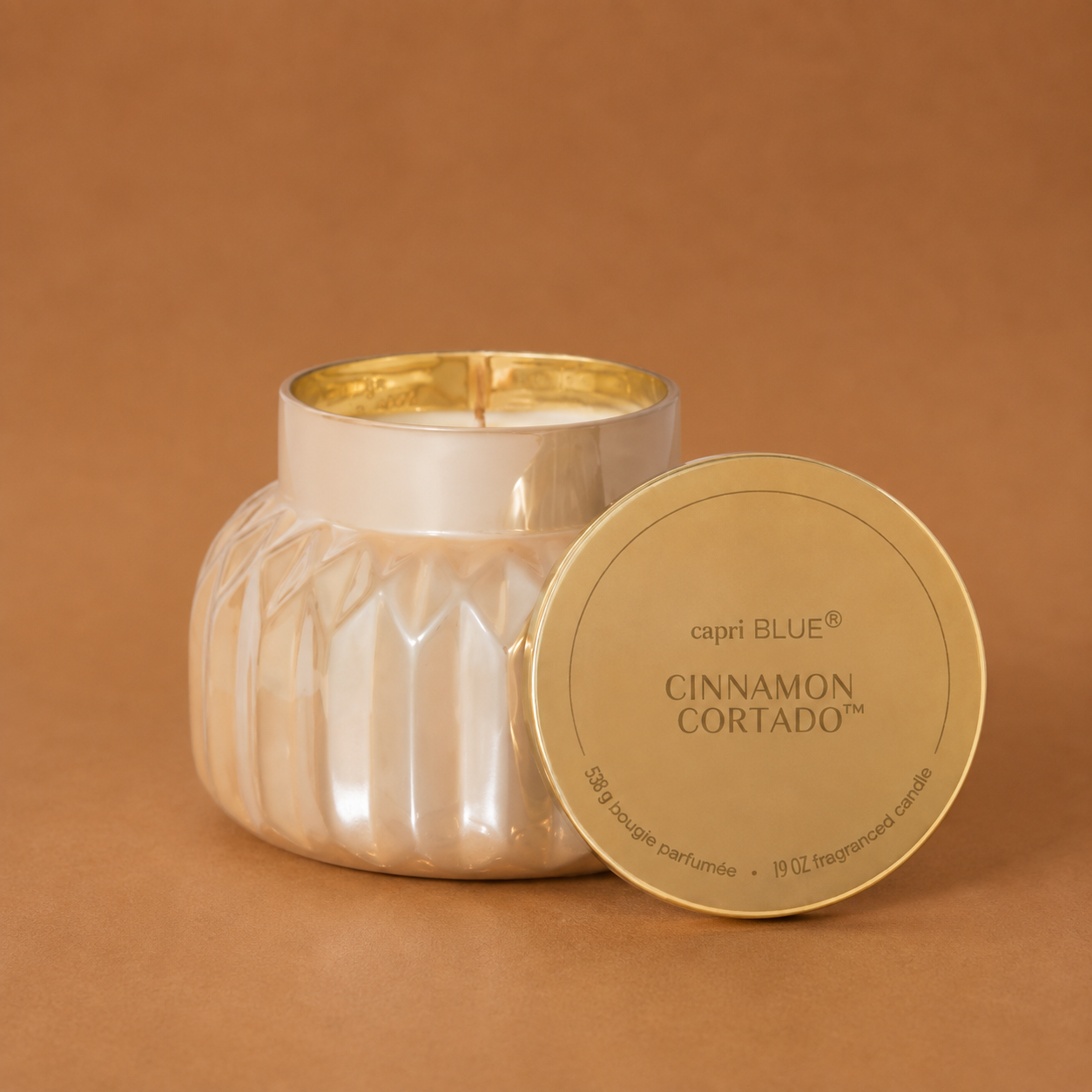 A silver faceted glass candle jar with its lid leaning against it on a warm brown background. The lid is gold-toned and printed with the brand name 'capri BLUE' and scent name 'CINNAMON CORTADO'.