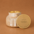 A silver faceted glass candle jar with its lid leaning against it on a warm brown background. The lid is gold-toned and printed with the brand name 'capri BLUE' and scent name 'CINNAMON CORTADO'.