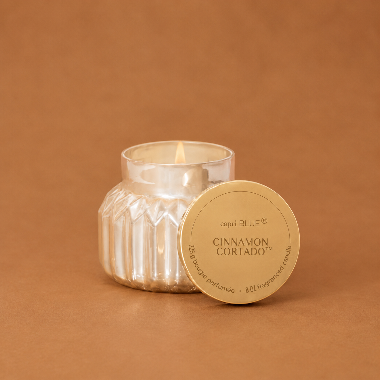 A lit scented candle in a ribbed glass jar with a round gold lid placed in front, set against a warm brown background. The lid reads 'capri BLUE' and 'CINNAMON, CORTADO'.
