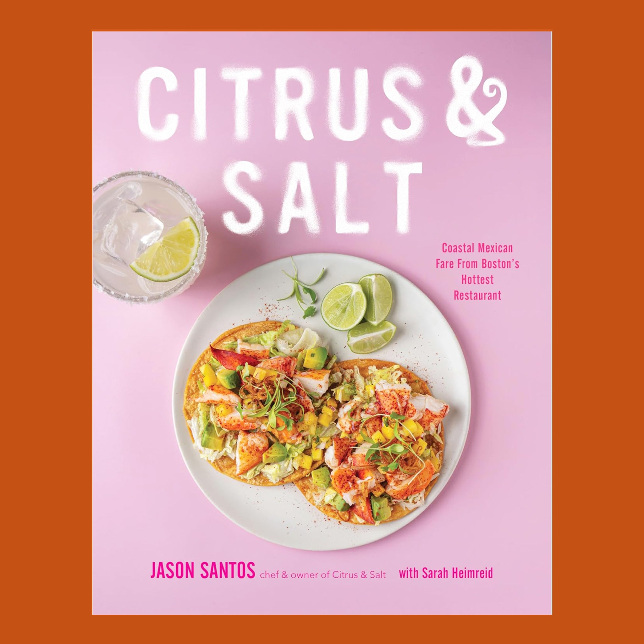 The cover of a pink cookbook with the title, "Citrus & Salt".  There are two shrimp tacos on the cover with limes and avocado.  The book is on an orange background.
