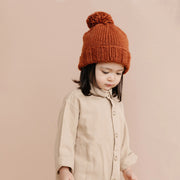 On a peachy background is a model wearing a reddish colored knit beanie with a pom pom on top.