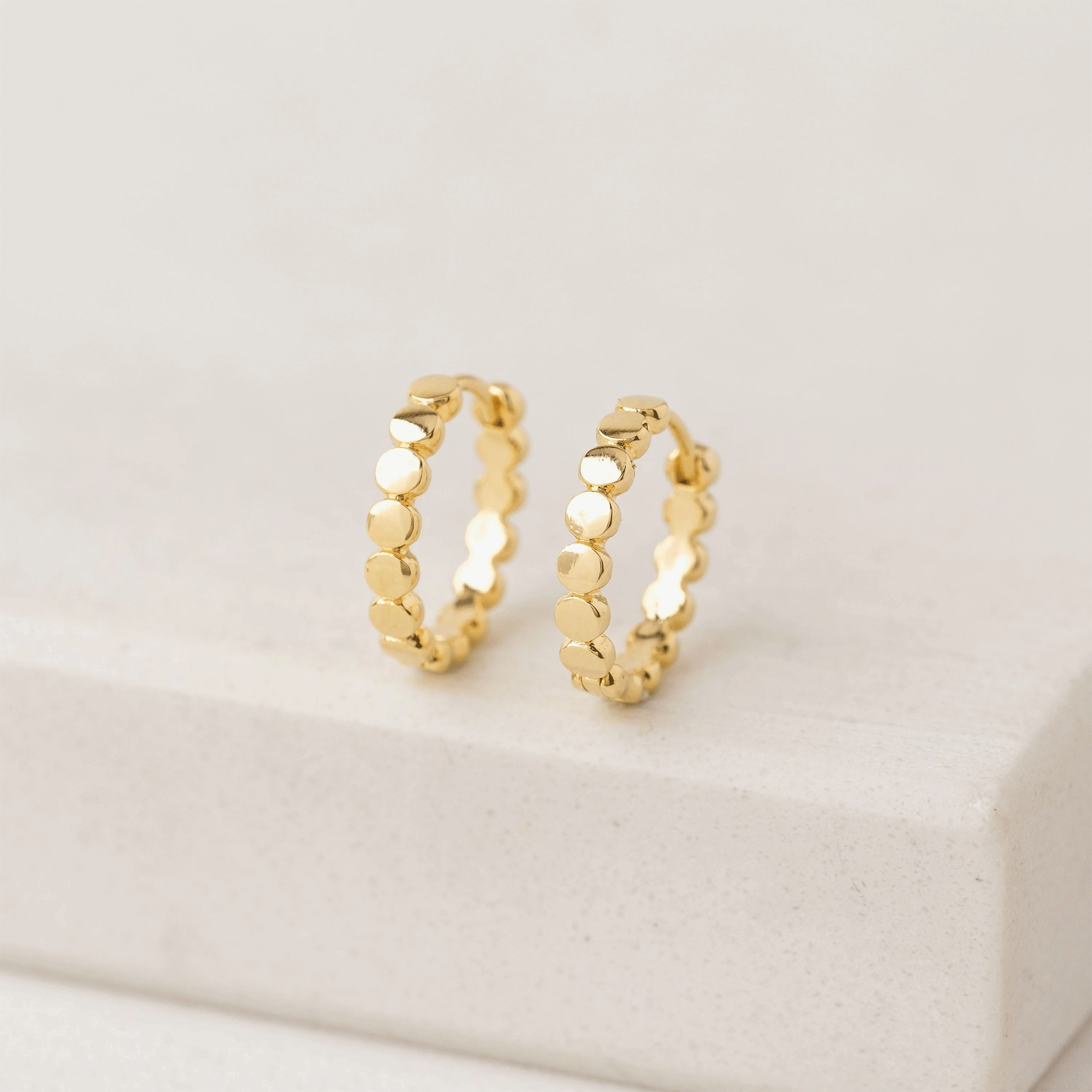 On a tan background is a pair of gold hoop earrings with a dainty circle detailing on the hoops. 