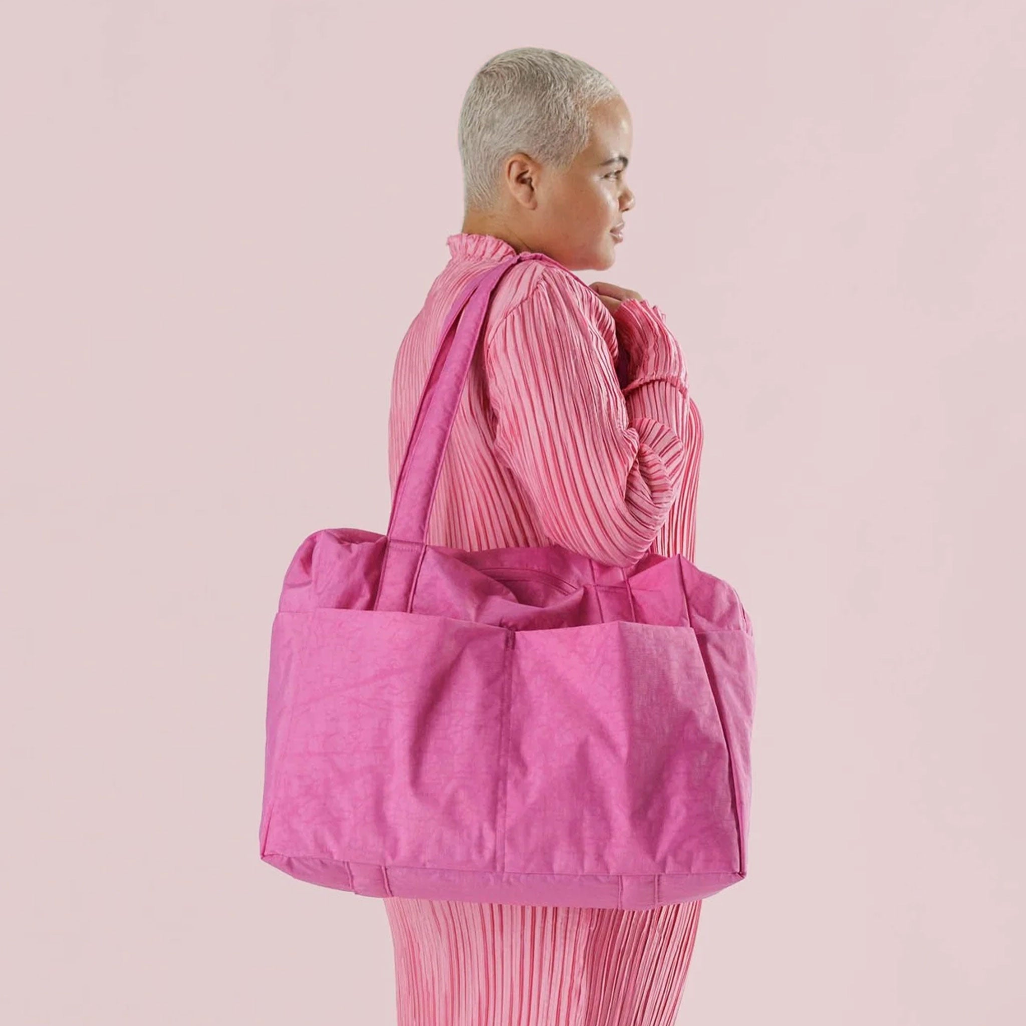On a light pink background is a bright pink tote bag with a shoulder strap and pockets.
