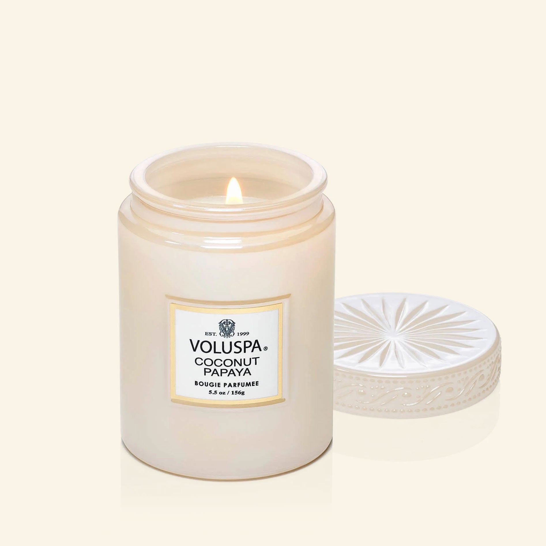 On a neutral background is a light pink glass jar candle with a white label with "Voluspa Coconut Papaya". 