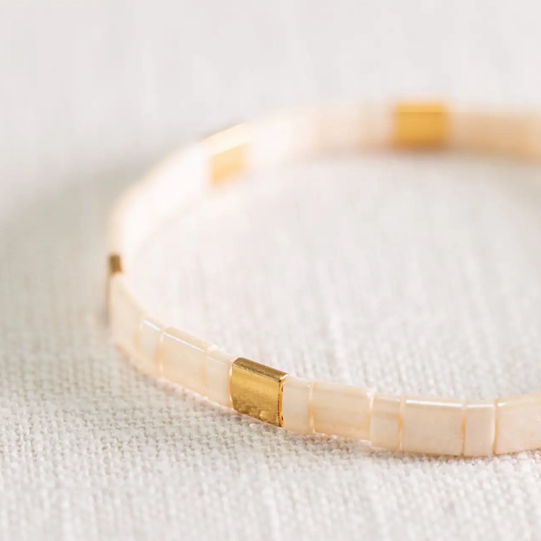 On a white background is a light tan and gold beaded bracelet. 